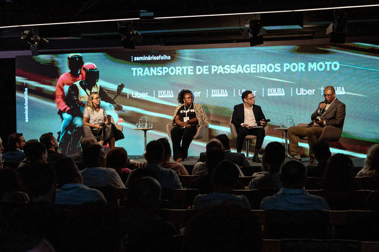 A imagem mostra um seminário com quatro pessoas sentadas em um palco， discutindo o tema 039;Transporte de Passageiros por Moto039;. Ao fundo， há uma tela com a imagem de motociclistas e o título do seminário em destaque. O público está visível， assistindo ao evento. Os participantes incluem uma mulher com cabelo longo e liso， uma mulher com cabelo crespo e uma camisa estampada， um homem com um terno claro e um homem com um terno escuro. Há copos de água sobre a mesa.
