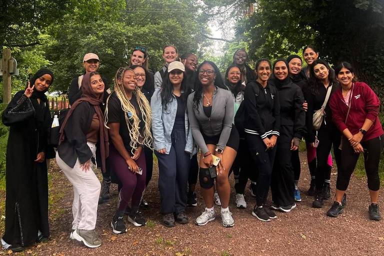 Um grupo de mulheres se reúne e posa para uma foto no caminho para uma área arborizada. Elas são de origens diferentes， algumas se agacham um pouco para a foto ou se inclinam， um casal faz um sinal positivo de V para a câmera. Atrás delas， há arbustos e árvores verdes com uma área aberta marrom sob seus pés