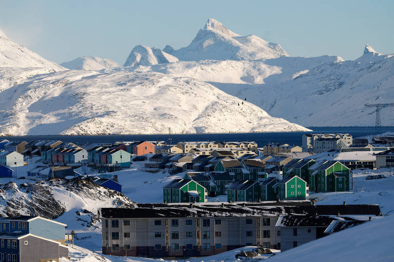 A imagem mostra uma paisagem coberta de neve， com montanhas ao fundo e um céu claro. Na parte inferior， há várias construções coloridas， predominantemente em tons de verde e azul， que contrastam com o branco da neve. O mar é visível entre as montanhas e as construções