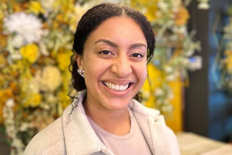 Uma mulher com cabelo preto preso para trás， usando um brinco na orelha direita， uma camisa roxa clara e uma jaqueta branca， está sorrindo em pé em frente a uma parede coberta de flores em um restaurante