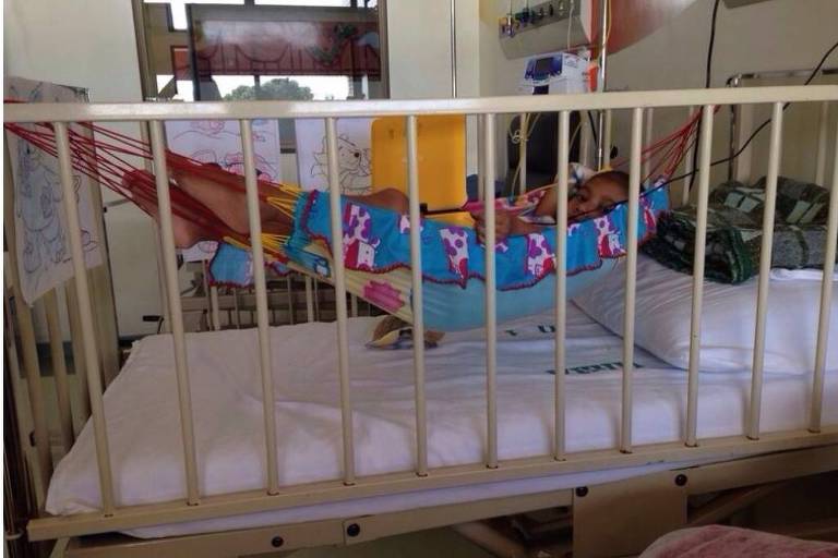 Uma criança está deitada em uma rede colorida dentro de um berço hospitalar. O berço é cercado por grades e a rede é decorada com desenhos de unicórnios. Ao fundo， há uma cama com lençóis brancos e alguns equipamentos médicos visíveis. A iluminação é natural， entrando pela janela.