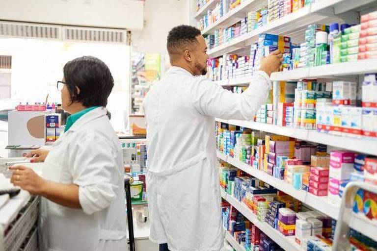 A imagem mostra uma farmácia com prateleiras cheias de medicamentos. Dois funcionários， um homem e uma mulher， estão organizando os produtos. O homem está em pé， alcançando uma prateleira alta， enquanto a mulher está em uma posição mais baixa， observando ou manipulando algo em uma mesa. Ambos estão usando jalecos brancos.
