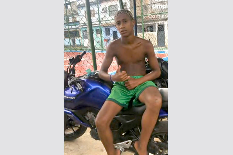 Um jovem está sentado em uma motocicleta azul. Ele está sem camisa e usa um short verde. O fundo mostra uma área urbana com edifícios e uma quadra de esportes.