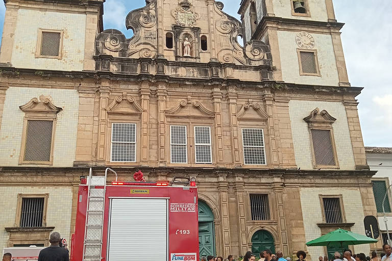 Foto colorida mostra igreja dourada ao fundo. Na frente do templo uma ambulância e diversas pessoas