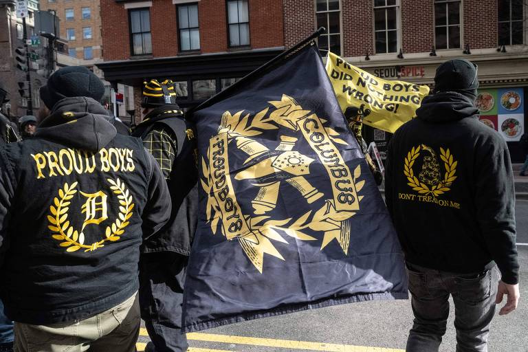 A imagem mostra um grupo de pessoas participando de uma manifestação. Algumas delas estão usando jaquetas pretas com emblemas dourados nas costas. Um dos manifestantes segura uma bandeira grande， também em preto e dourado， que apresenta um símbolo central. Ao fundo， há edifícios e uma faixa amarela visível