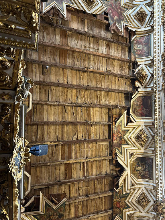 Imagem do teto de uma igreja， com painéis de madeira dispostos em linhas verticais. O teto apresenta detalhes decorativos em forma de estrelas e ornamentos dourados. A iluminação é suave， destacando a textura da madeira e os elementos decorativos.