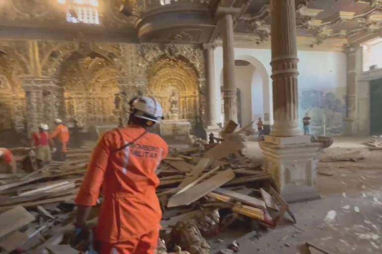 A imagem mostra um interior de um edifício danificado， com destroços de madeira espalhados pelo chão. Há várias pessoas vestindo uniformes de resgate， algumas com capacetes， trabalhando na área. O ambiente apresenta paredes ornamentadas e colunas， mas está em estado de desordem devido aos danos. A luz natural entra pelas janelas， iluminando a cena.