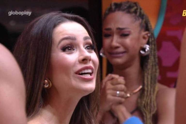A imagem mostra duas mulheres em um ambiente de programa de televisão. A mulher à esquerda, com cabelo longo e solto, está sorrindo e parece estar falando animadamente. A mulher à direita, com cabelo trançado e expressão de choro, está com as mãos unidas em frente ao rosto, demonstrando emoção. Ao fundo, é possível ver uma parede colorida e parte de outra pessoa.
