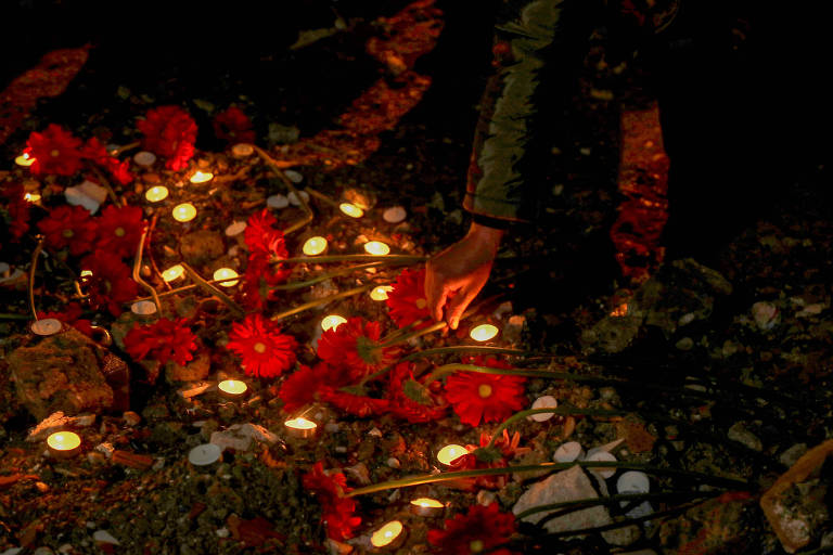 A imagem mostra uma cena noturna onde há várias velas acesas ao redor de flores vermelhas. Uma mão está posicionada para acender uma das velas, enquanto as flores estão dispostas no chão, criando um ambiente de homenagem ou lembrança.