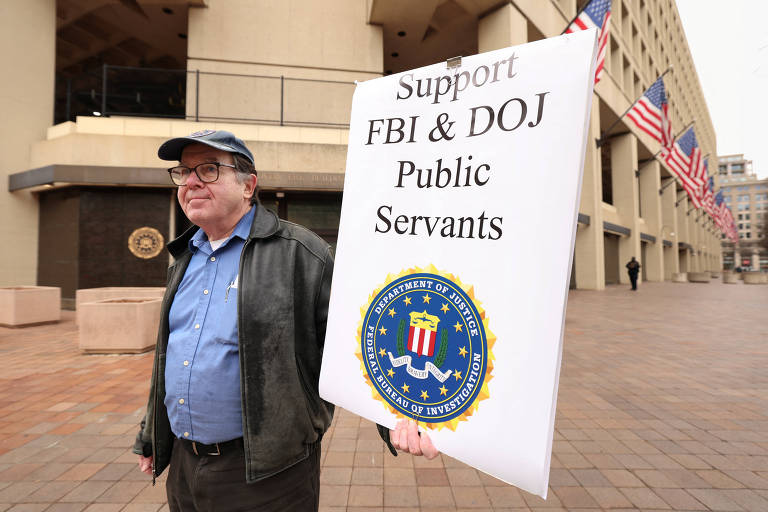 Um homem está em pé segurando um cartaz que diz 039;Support FBI amp; DOJ Public Servants039;. Ele usa um boné e óculos， e está vestido com uma camisa azul e um casaco escuro. Ao fundo， há um edifício com bandeiras dos Estados Unidos.