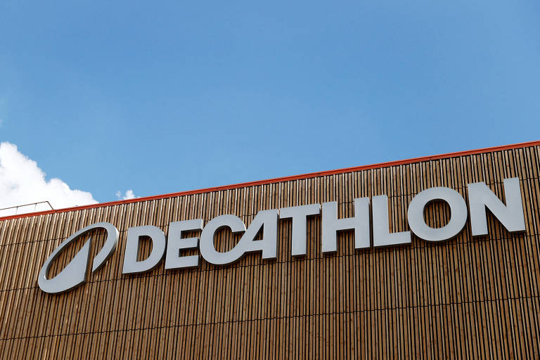 A imagem mostra a fachada de uma loja da Decathlon， com o logotipo da marca em letras grandes e brancas. O fundo é um céu azul com algumas nuvens. A estrutura da loja é de madeira， com um design moderno.