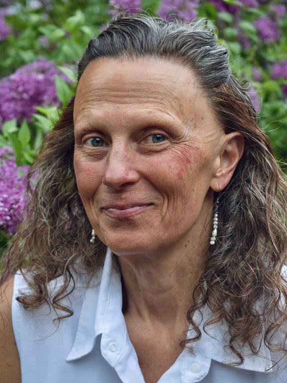 A imagem mostra uma mulher com cabelo cacheado e solto， usando uma blusa branca. Ela está sorrindo levemente e tem brincos de pérola. Ao fundo， há flores lilases e folhagens verdes， criando um ambiente natural e vibrante.