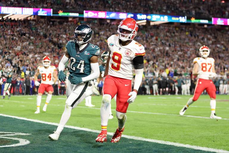 A imagem mostra um jogador da equipe Philadelphia Eagles， vestido com um uniforme verde e o número 24， correndo em direção à end zone. Um jogador da equipe Kansas City Chiefs， vestido com um uniforme vermelho e o número 9， está próximo a ele. Ao fundo， há uma multidão assistindo ao jogo， com outros jogadores visíveis em campo.
