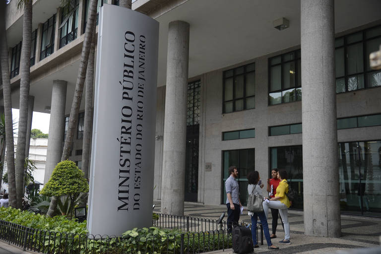 A imagem mostra a fachada de um edifício moderno do Ministério Público. Há um grande letreiro vertical com a inscrição 039;MINISTÉRIO PÚBLICO039; em letras maiúsculas. Na frente do edifício， algumas pessoas estão conversando， e há uma área verde com plantas ao redor.