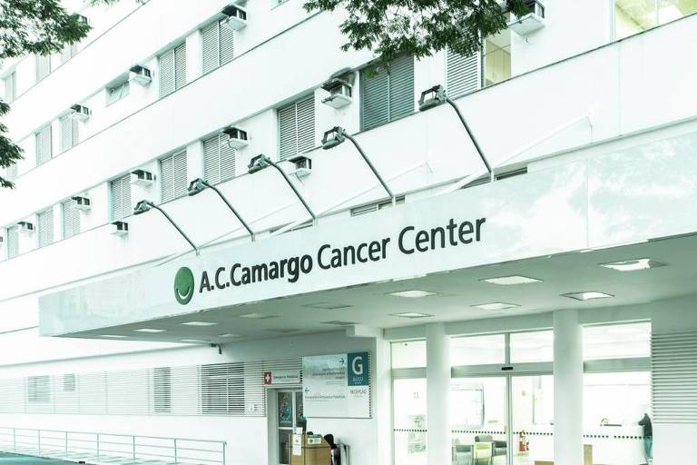 Imagem do exterior do A.C. Camargo Cancer Center， um edifício de vários andares com uma fachada clara. A entrada principal é visível， com portas de vidro e uma área de estacionamento em frente. Algumas árvores estão presentes ao redor do edifício.