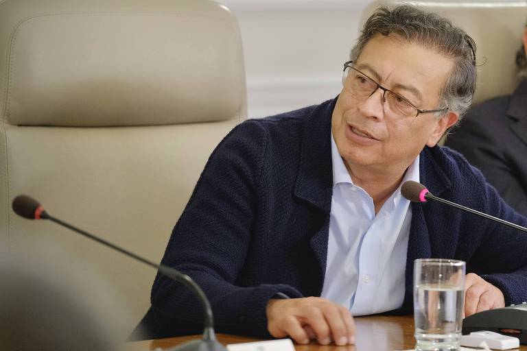 A imagem mostra uma reunião em uma mesa de conferência. Um homem com óculos e cabelo ondulado， vestido com um suéter escuro， está falando. Ao fundo， outro homem com cabelo grisalho e barba está ouvindo atentamente. Há copos de água e documentos sobre a mesa.