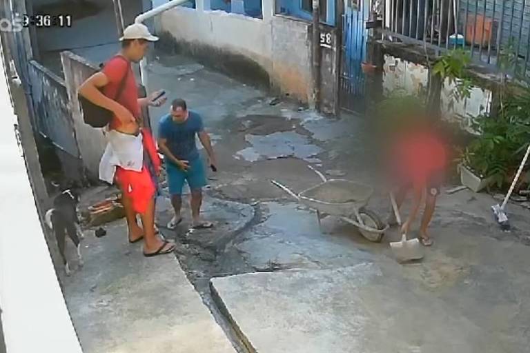 A imagem mostra uma cena capturada por uma câmera de segurança em uma rua. Há três pessoas visíveis: um homem em pé à esquerda， vestindo uma camiseta vermelha e shorts， segurando uma bolsa; um homem ao centro， com uma camiseta azul; e uma pessoa à direita， parcialmente desfocada. O ambiente é uma rua de uma área urbana， com casas ao fundo e um piso de concreto. Algumas plantas estão visíveis ao lado.