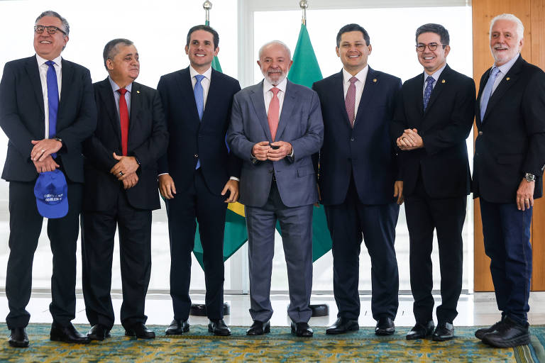 A imagem mostra um grupo de seis homens em um ambiente interno， posando para a foto. Eles estão vestidos formalmente， com ternos e gravatas. Ao fundo， há bandeiras do Brasil. O homem no centro parece ser o foco principal da imagem， segurando algo com as mãos. Os outros homens estão dispostos ao seu redor， sorrindo.
