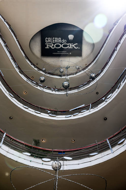 A imagem mostra a parte interna da Galeria do Rock, com uma vista de baixo para cima. O teto apresenta um grande letreiro com a inscrição 'GALERIA do ROCK'. O espaço é circular, com várias varandas e uma estrutura de iluminação pendurada no centro, composta por esferas metálicas. As paredes são de um tom claro e há detalhes em vermelho nas varandas.