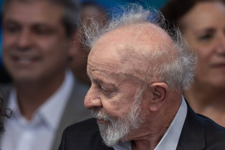 A imagem mostra um homem idoso com cabelo grisalho e bagunçado, usando um terno escuro e uma camisa clara. Ele está de perfil, olhando para a direita, enquanto ao fundo há outras pessoas sorrindo, algumas delas com roupas formais. O ambiente parece ser um evento social.