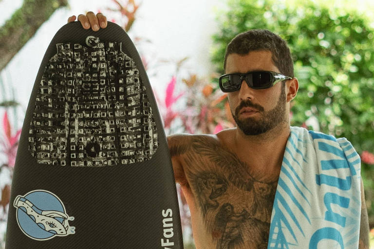 Um homem musculoso, sem camisa, está posando com uma prancha de surf preta em uma mão e uma toalha azul e branca na outra. Ele usa óculos escuros e tem tatuagens visíveis no braço. Ao fundo, há plantas verdes e um ambiente externo. A prancha possui adesivos com os textos 'OnlyFans' e um logotipo de um golfinho. A toalha também exibe o logotipo 'OnlyFans'.