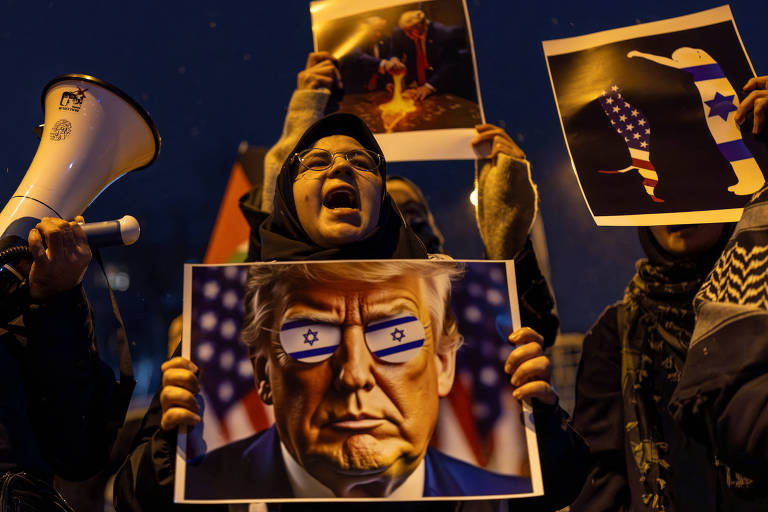 A imagem mostra um grupo de pessoas em um protesto à noite. Uma mulher， usando um lenço na cabeça， segura um cartaz com a imagem de um homem com óculos que têm a bandeira de Israel. Ao fundo， outras pessoas seguram cartazes e um megafone. O ambiente é escuro， com algumas luzes visíveis.