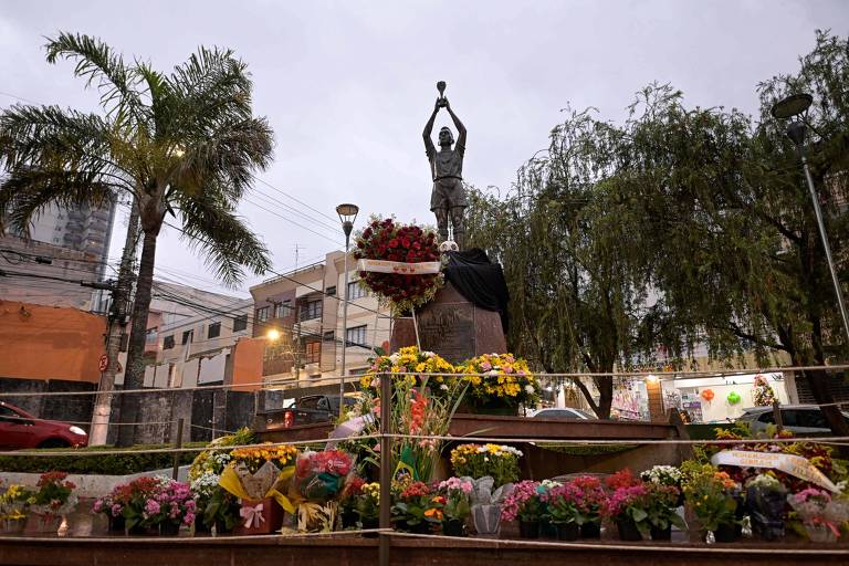 A imagem mostra uma estátua de um atleta segurando uma bola acima da cabeça， cercada por flores coloridas e arranjos florais. Ao fundo， há prédios e árvores， com um céu nublado. A estátua está em uma área pública， com iluminação visível e um ambiente urbano.
