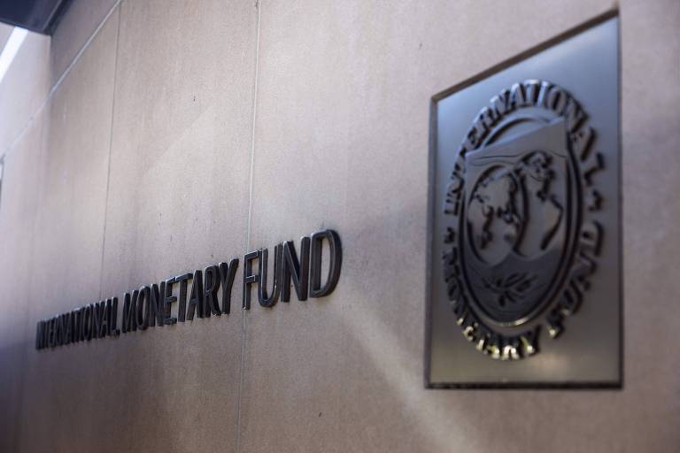 A imagem mostra a fachada do Fundo Monetário Internacional (FMI)， com o nome 039;INTERNATIONAL MONETARY FUND039; em letras grandes e em destaque. Ao lado， há um emblema do FMI， que apresenta um desenho estilizado de um globo terrestre e figuras humanas. O fundo é de uma parede clara.
