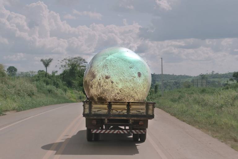 A imagem mostra um caminhão transportando um grande objeto esférico que parece ter uma superfície semelhante à da lua, com crateras e uma coloração que varia entre tons de marrom e verde. O caminhão está em uma estrada rural, cercado por vegetação e sob um céu parcialmente nublado, com nuvens brancas e algumas árvores ao fundo.