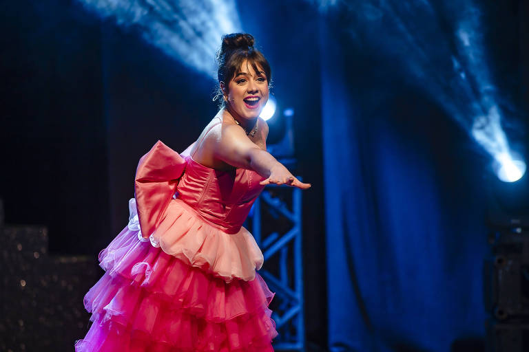 Uma mulher está se apresentando em um palco, usando um vestido rosa com camadas e um laço grande nas costas. Ela está sorrindo e estendendo a mão em direção ao público, com luzes de palco ao fundo criando um ambiente vibrante.