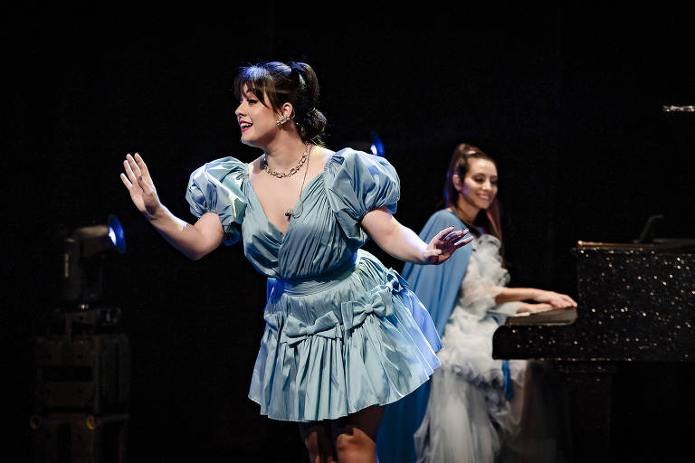 Uma mulher em um vestido azul claro está se apresentando no palco, gesticulando com as mãos. Ao fundo, outra mulher está sentada ao piano, vestindo um traje elegante com detalhes em branco. O ambiente é escuro, com luzes focadas nas artistas.