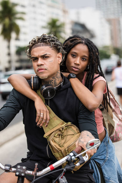 Um homem e uma mulher estão em uma bicicleta em uma área urbana. O homem, com cabelo encaracolado e tatuagens visíveis, usa uma camiseta de manga longa e fones de ouvido. Ele segura o guidão da bicicleta verde. A mulher, com cabelo trançado, está atrás dele, usando uma blusa rosa. Ao fundo, há prédios altos e palmeiras, e um táxi amarelo passa pela rua