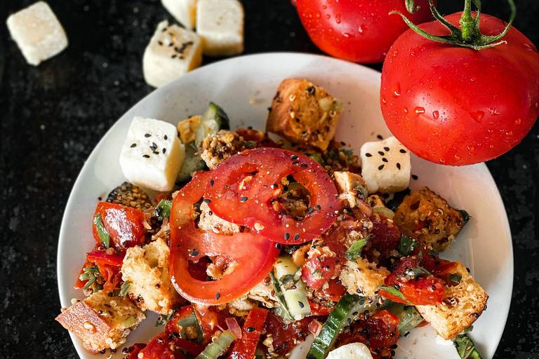 Um prato branco com uma salada que contém pedaços de pão， tomate fatiado， cubos de queijo e vegetais. Ao redor do prato， há tomates inteiros e cubos de queijo adicionais. O fundo é escuro