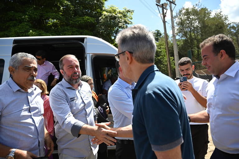 A imagem mostra um grupo de homens em um ambiente externo， próximo a um veículo de transporte. Alguns homens estão se cumprimentando， enquanto outros observam. Ao fundo， há árvores e postes de eletricidade. O clima parece ensolarado.
