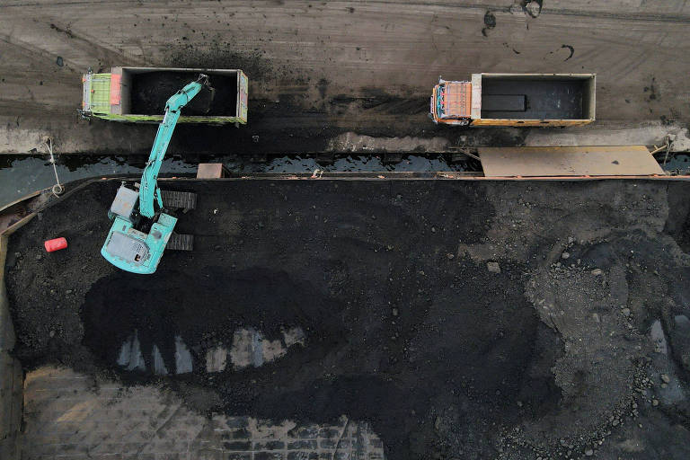 A imagem mostra uma vista aérea de um local de construção. Há duas caçambas de caminhão posicionadas em um terreno coberto de asfalto. Um equipamento de escavação， pintado de verde e azul， está em operação， aparentemente removendo material do solo. O chão apresenta manchas de asfalto e algumas marcas visíveis.
