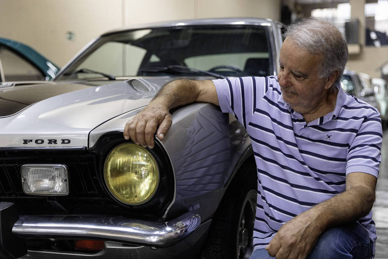 Um homem de cabelos grisalhos, vestido com uma camisa listrada em tons de azul e branco, está sentado ao lado de um carro Ford. Ele apoia o braço sobre o capô do carro, olhando para a frente com uma expressão pensativa. O carro é de cor cinza com detalhes em preto e possui um farol amarelo. O ambiente ao fundo é um espaço fechado, possivelmente uma garagem ou um museu de automóveis.