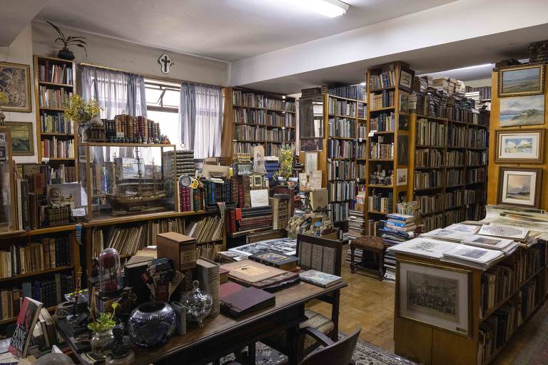 A imagem mostra uma biblioteca repleta de estantes cheias de livros organizados. Há mesas de madeira com objetos decorativos e livros abertos. As paredes são adornadas com quadros e há janelas com cortinas. O ambiente é bem iluminado, com uma atmosfera acolhedora e intelectual.