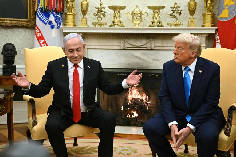 A imagem mostra dois homens brancos sentados em cadeiras no Salão Oval da Casa Branca. À esquerda， Netanyahu， um homem de cabelo grisalho e terno escuro com uma gravata vermelha， gesticulando com as mãos. À direita， Trump， um homem de cabelo loiro e terno escuro com uma gravata azul， olhando para frente. Ao fundo， uma lareira acesa e prateleiras com troféus e bandeiras.