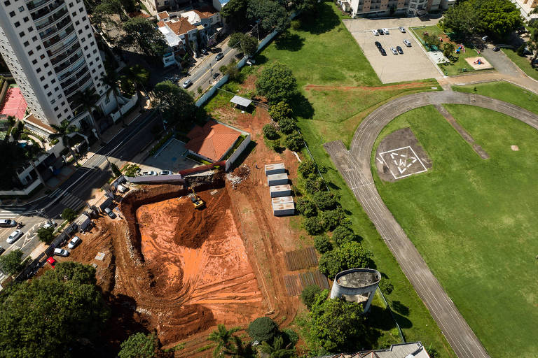 A imagem mostra uma vista aérea de uma área em construção， com um grande buraco escavado no solo， cercado por terra vermelha. À esquerda， há um edifício alto e， na parte inferior， veículos estacionados ao longo da rua. À direita， uma área verde com um campo e uma pista de corrida， além de um espaço de estacionamento. Algumas árvores estão visíveis ao redor da área.