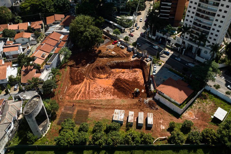 Imagem aérea de uma área de construção， mostrando um terreno com solo exposto e marcas de máquinas. À esquerda， há casas com telhados vermelhos e árvores. À direita， um edifício alto e uma rua com carros. No fundo， há um tanque de água ou estrutura circular e contêineres de armazenamento.