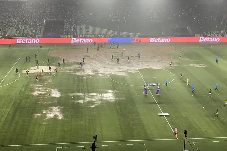 A imagem mostra um campo de futebol com áreas de grama danificada e lama， onde jogadores estão em campo. O ambiente parece nebuloso， com uma iluminação intensa ao fundo e uma grande quantidade de pessoas ao redor do campo. As linhas do campo são visíveis， mas a condição do gramado é precária.
