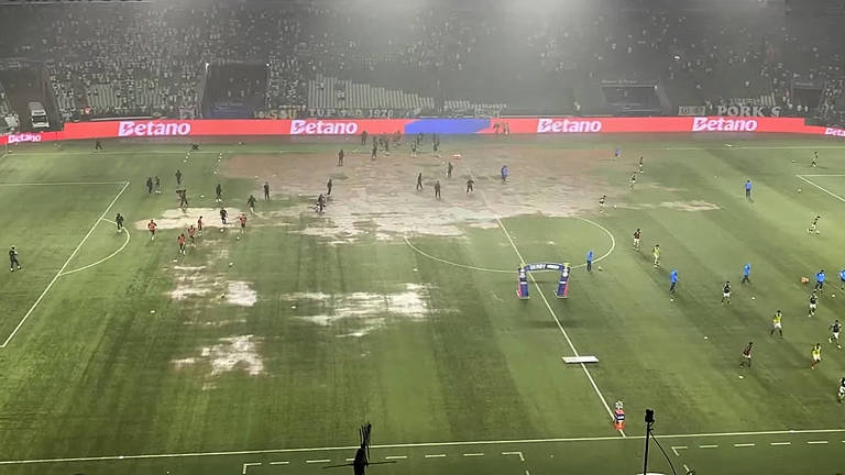 A imagem mostra um campo de futebol com áreas de grama danificada e lama， onde jogadores estão em campo. O ambiente parece nebuloso， com uma iluminação intensa ao fundo e uma grande quantidade de pessoas ao redor do campo. As linhas do campo são visíveis， mas a condição do gramado é precária.