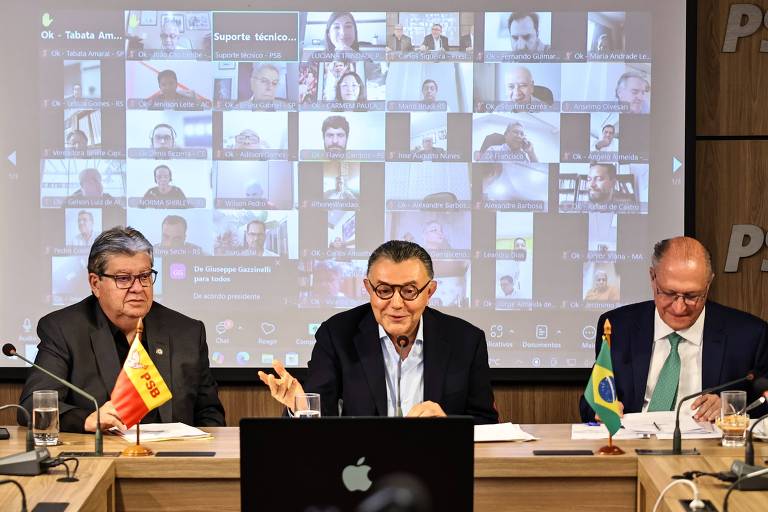 A imagem mostra uma reunião virtual com três homens sentados à mesa. O homem à esquerda está com uma bandeira， o do meio está gesticulando e o da direita está com uma gravata. Ao fundo， há uma tela exibindo várias janelas de participantes em videoconferência.