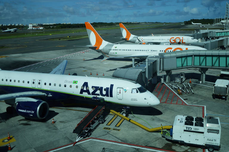 A imagem mostra uma área de embarque em um aeroporto， com dois aviões da companhia aérea Gol e um avião da companhia aérea Azul. O céu está parcialmente nublado e há uma estrutura de terminal ao fundo.
