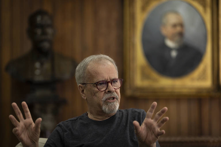 Ricardo Teixeira, um homem de cabelos grisalhos e barba, usando óculos e uma camiseta escura, está sentado e gesticulando enquanto fala. Ao fundo, há um busto de uma figura masculina e um retrato emoldurado de um homem com barba e gravata. O ambiente é de madeira escura, sugerindo um espaço formal ou histórico.