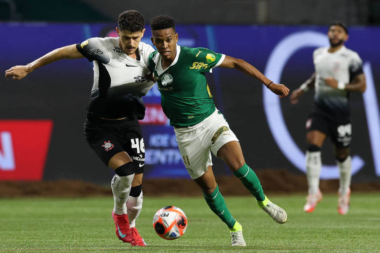 Dois jogadores de futebol estão em uma disputa pela bola em um campo. Um jogador veste uma camisa preta e branca， enquanto o outro está vestido com uma camisa verde. O jogador da camisa verde está tentando driblar o adversário， que está se aproximando para desarmá-lo. Ao fundo， é possível ver outros jogadores e uma tela publicitária.
