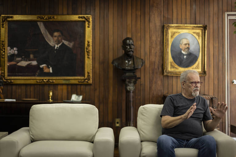 A imagem mostra um homem sentado em um sofá branco em um ambiente com paredes de madeira. Ele está usando uma camiseta cinza e óculos. O homem gesticula com as mãos enquanto fala. Ao fundo, há um busto e duas pinturas emolduradas, uma delas retratando um homem em traje formal e a outra um homem mais velho. O ambiente parece ser um escritório ou sala de reuniões.