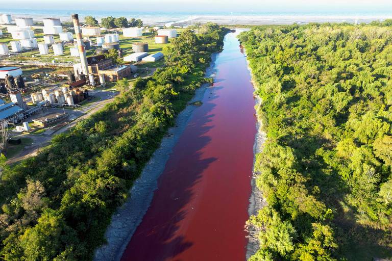 Imagem aérea de um rio com coloração vermelha， cercado por vegetação densa de ambos os lados. Ao fundo， há tanques de armazenamento e uma chaminé de uma indústria， indicando a presença de atividade industrial nas proximidades.