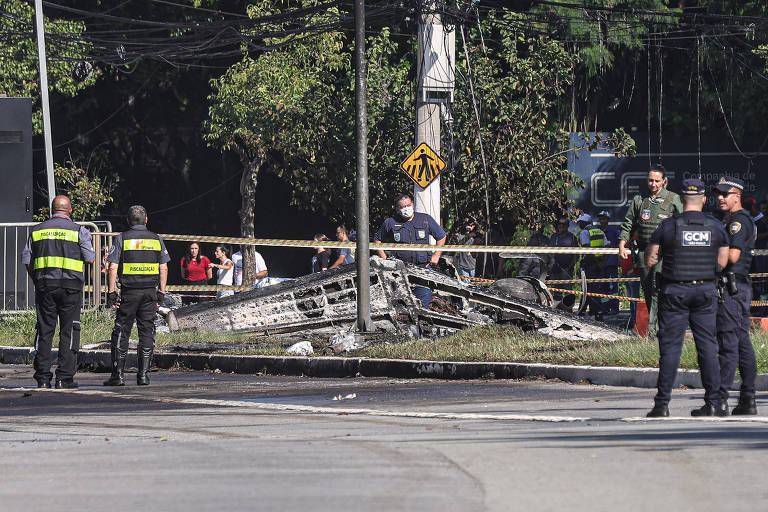 A imagem mostra o local de um acidente de veículo, onde um carro está completamente destruído e queimado, próximo a um poste de eletricidade. Há várias pessoas, incluindo policiais e membros de equipes de emergência, ao redor do local, com algumas pessoas observando ao fundo. O ambiente é urbano, com árvores e sinais de trânsito visíveis.