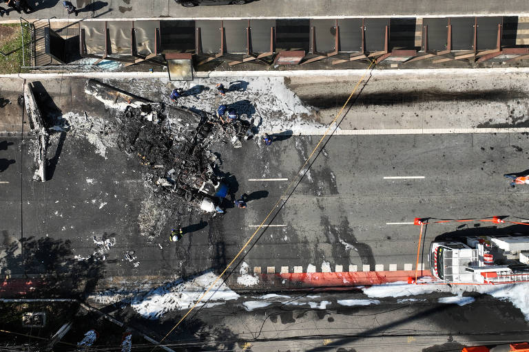 A imagem mostra uma vista aérea de uma estrada onde ocorreu um acidente. Há destroços queimados e uma área coberta de espuma branca， possivelmente de um combate a incêndio. Veículos de emergência estão presentes na cena， e a estrada parece estar bloqueada.